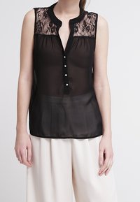 Blouse noire sans manches avec une superposition de dentelle sur les épaules, col rond et boutonnage sur le devant. Tissu transparent avec un détail d'ourlet plissé.