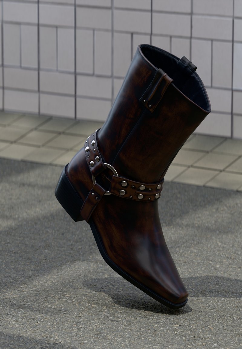 Καφέ σκούρο δερμάτινο μποτάκι μέχρι τον αστράγαλο με ασημένια τρουκς και λεπτομέρεια με λουράκι, εμφανιζόμενο μπροστά σε τοίχο με πλακάκια και φόντο από σκυρόδεμα.