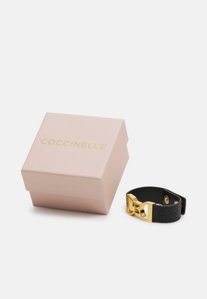 Bracelet en cuir noir avec un détail de boucle en or, placé à côté d'une boîte rose pâle portant le nom de la marque "Coccinelle" en lettres dorées.