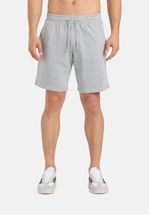 Uomo con pantaloncini elasticizzati grigio chiaro con coulisse in vita e sneakers bianche con strisce nere, in piedi davanti a uno sfondo neutro.