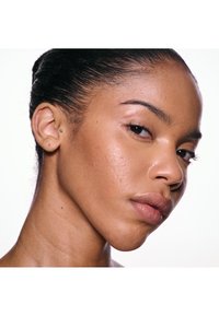 Gros plan sur une femme au teint lisse, maquillage naturel, cheveux foncés tirés en arrière, une petite piercing à l'oreille et une expression neutre, légèrement tournée sur le côté.