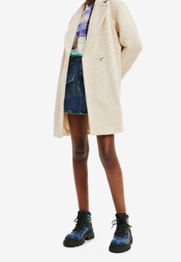 Manteau long texturé beige sur un t-shirt graphique, associé à une jupe en jean et des bottines à lacets bleu-noir avec une semelle  épaisse.