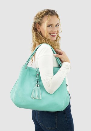 Vrouw met krullend blond haar, gekleed in een witte lange mouwen top en blauwe jeans, die glimlacht en een grote lichtblauwe leren handtas vasthoudt.