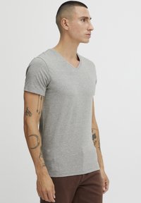 Siva v-neck majica iz mehkih materialov, prilegajoče oblikovanje, kratke rokave, z rahlo teksturo. Obuta z rjavimi hlačami in vidnimi tetovažami na rokah.