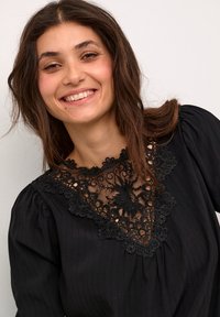 Blusa nera con maniche a sbuffo, scollatura in pizzo ricamato trasparente con motivi floreali e un design in tessuto texturizzato.