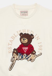 Εκρού φούτερ με κεντητό αρκουδάκι που φοράει κόκκινο πουλόβερ και τρίγωνο λογότυπο, με την επιγραφή "Ιδρύθηκε το 1981" σε μπεζ χρώμα.