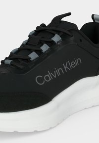 Baskets noires Calvin Klein avec œillets gris, lacets noirs et semelle blanche épaisse sur un fond clair.