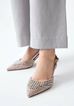 Piedi che indossano pantaloni beige chiaro e décolleté slingback a punta ricoperte di strass scintillanti su sfondo bianco.