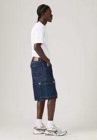 Șorturi cargo din denim de culoare albastru închis, cu buzunare aplicate și închideri cu capse, asortate cu un tricou alb și adidași gri cu accente metalice.