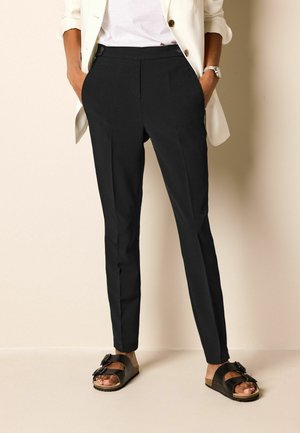 Persona con pantalones negros a medida, camisa blanca, blazer crema, sandalias negras, de pie con las manos en los bolsillos frente a un fondo liso.