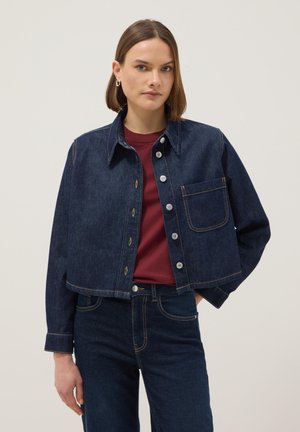 Mujer con cabello corto y castaño vestida con chaqueta de mezclilla oscura y jeans sobre una camisa marrón oscuro, de pie frente a un fondo claro y liso.