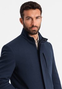 Giacca blu navy con colletto alto, tessuto strutturato e tasca laterale con zip. Design aderente con una silhouette elegante e dettagli moderni.