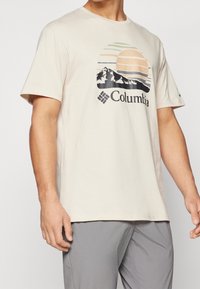 Beige tričKo s krátkým rukávem a kulatým výstřihem. Na přední straně je grafika hor a slunce s logem "Columbia" pod ní. Měkký bavlněný materiál.