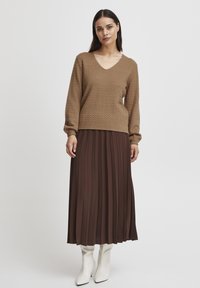 Pull en tricot marron avec un col en V, associé à une longue jupe plissée marron et des bottes blanches hautes jusqu'aux genoux. Tissu texturé sur le pull.