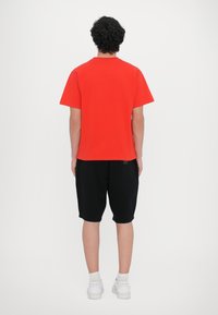 Rotes T-Shirt mit kurzen Ärmeln, Rückansicht, kombiniert mit schwarzen knielangen Shorts und weißen Sneakers, vor einem schlichten weißen Hintergrund präsentiert.