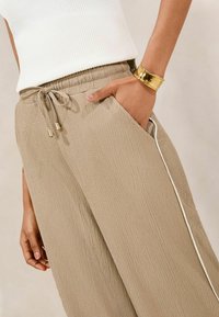 Béžové textured kalhoty s elastickým pasem, šňůrkou s kovovými konci a bočními kapsami. Ozdobené bílým pruhem po straně.