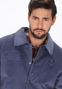 Mørkeblå corduroyjakke med knaplukning, spids krave, broderet ankerlogo og dekorativ syning på brystet.