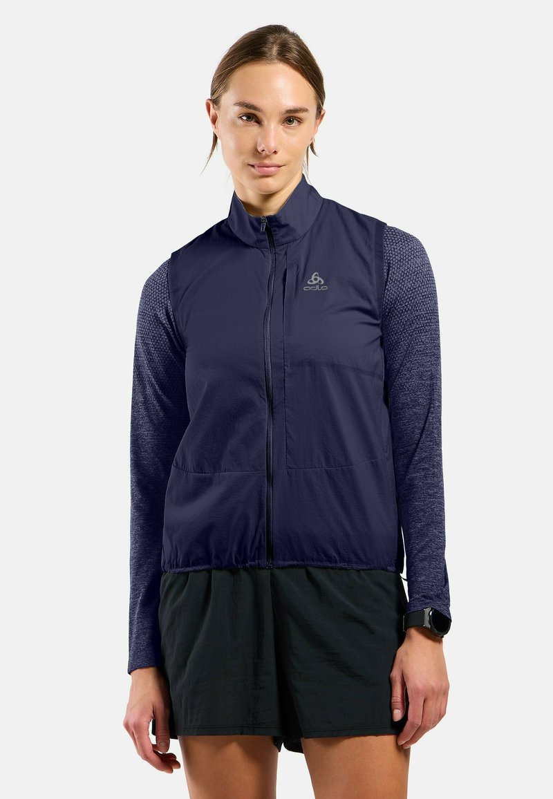 Marineblauer leichter Weste mit hohem Kragen, Frontreißverschluss, zwei Taschen und strukturierten langen Ärmeln. Getragen über schwarzen Shorts.