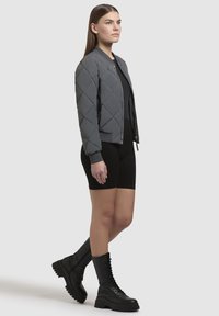 Grijze gewatteerde bomberjack met een rits aan de voorkant, gecombineerd met zwarte biker shorts en zwarte veterschoenen voor combat. Gladde, gevoerde textuur.