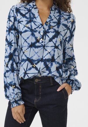 Niebieska bluzka tie-dye ze złotymi kwiatowymi akcentami, zapinany kołnierzyk, długie bufiaste rękawy z elastycznymi mankietami, zestawiona z ciemnoniebieskimi dżinsami.