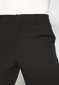 Zbliżenie na osobę noszącą czarne dopasowane spodnie z szlufkami na pasek i bocznymi kieszeniami, w połączeniu z białą, wpuszczoną do środka bluzką.