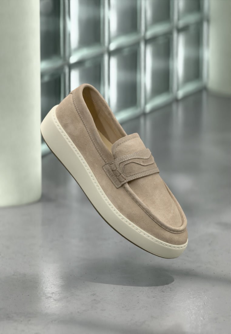 Mocassin en daim beige avec une semelle en caoutchouc blanc flottant au-dessus d'un sol en béton poli près d'un mur en blocs de verre.