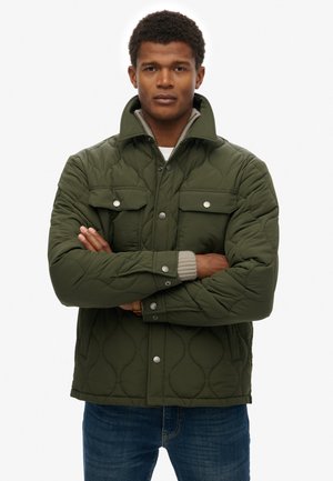 Giovane uomo con le braccia incrociate che indossa una giacca trapuntata verde con bottoni sopra a un maglione chiaro e jeans blu.