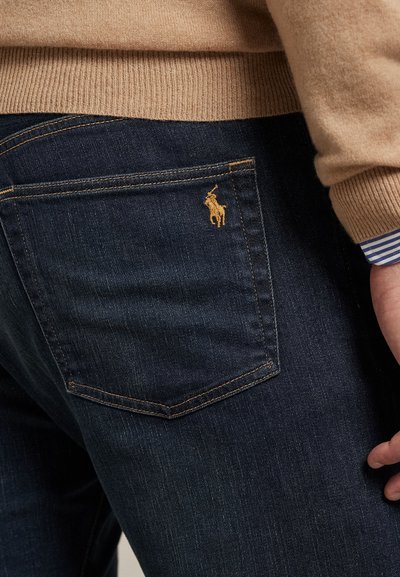 Temno modre denim kavbojke z teksturirano površino, na zadnjem žepu s rumenim vezenim logotipom in kontrastnim rumenim šivom.