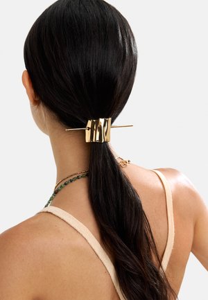 Widok z tyłu na kobietę z ciemnymi włosami spiętymi nisko w kucyk, zabezpieczonym dużą, błyszczącą złotą spinką do włosów z pałeczkowym detalem.