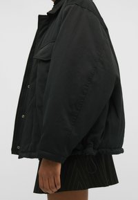 Sort oversized jakke med knapper foran, to brystlommer, elastiske manchetter og en justerbar kant, der har en glat, mat overflade.