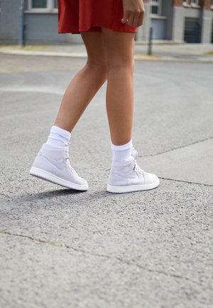Jambes d'une personne marchant sur le trottoir, portant des baskets blanches montantes, des chaussettes blanches et une jupe rouge.