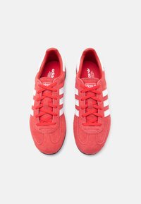 Röda sneakers i mocka med vita ränder, med rundad tå och texturerad sula. Skosnören och innerfoder är också röda.