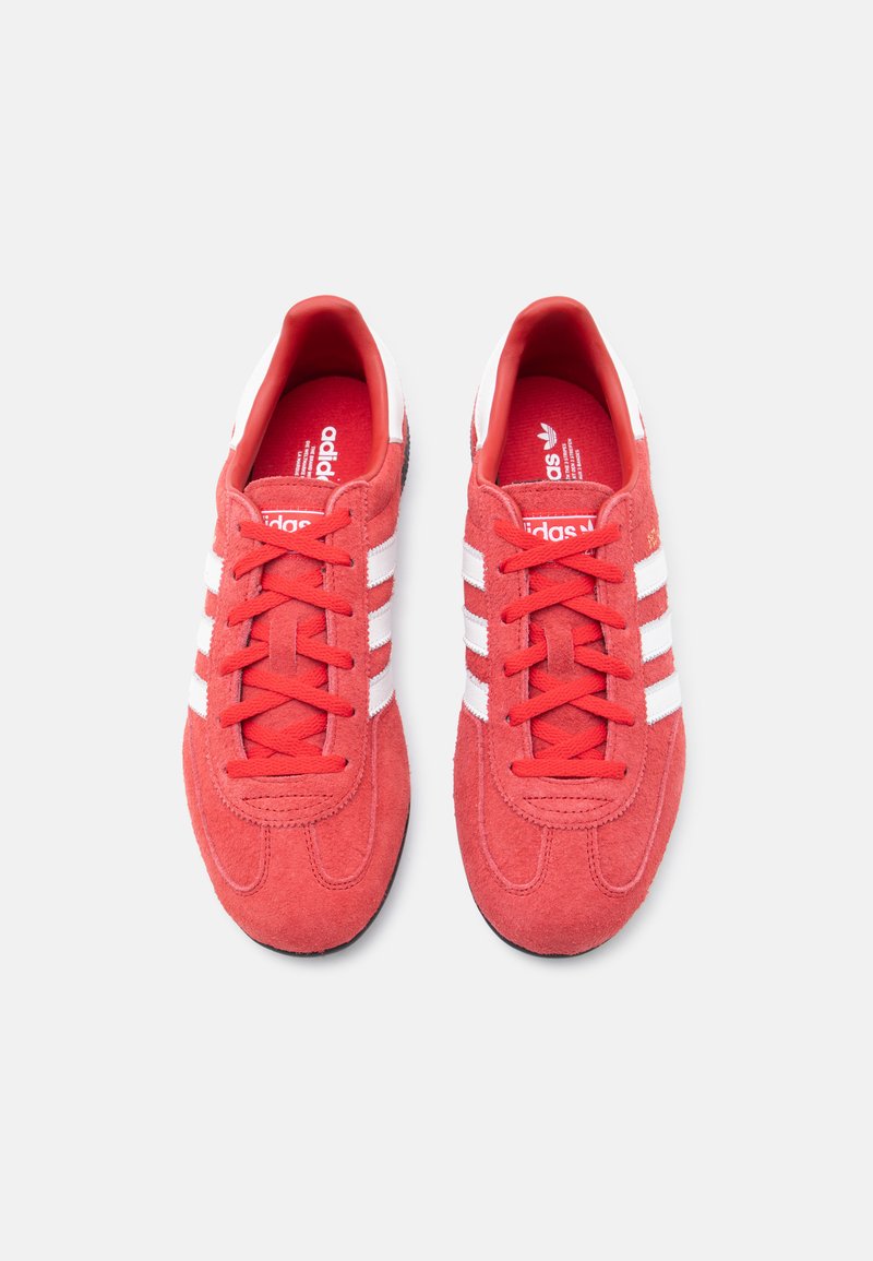 Red suede sneakers with white stripes, featuring a rounded toe and a textured sole. Laces and inner lining are also red.