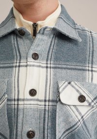 Giacca a quadri con tonalità di blu, bianco e grigio; realizzata in un tessuto con texture a spina di pesce; presenta bottoni scuri e due tasche frontali.