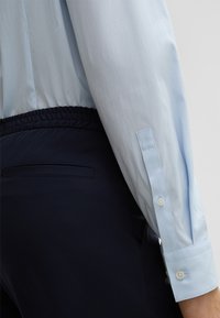 Lichtblauwe lange mouw shirt met knoopmanchetten, naast marineblauwe broek met elastische tailleband en een achterzakdetail.