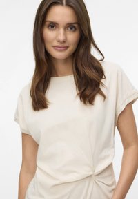 Camiseta de algodón color crema con mangas cortas enrolladas, detalle de nudo en la parte delantera, textura suave y corte relajado.