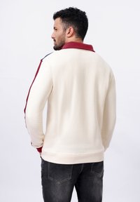 Polo shirt à manches longues crème avec un col bordeaux et des rayures latérales bleues, associé à un jean foncé. Texture douce avec une coupe décontractée.