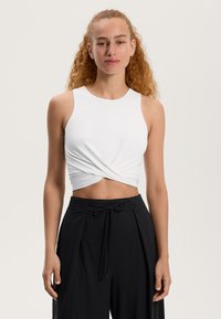 Bela skrajšana majica brez rokavov z zavitim sprednjim delom, v kombinaciji z visokim pasom črnimi hlačami, ki imajo zavezan pas in gubano detajl.