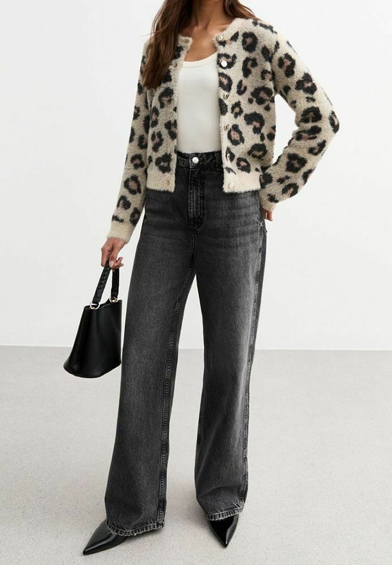 Femme portant un cardigan imprimé léopard, un haut blanc, un jean noir à jambes larges, des chaussures noires pointues et tenant un petit sac à main noir.
