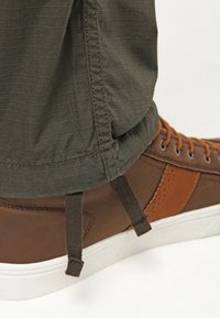 Brązowy skórzany sneaker z tekstylnymi akcentami, białą podeszwą i brązowymi sznurowadłami. Oliwkowozielone spodnie z tkaniny z wzorem kratki i ściągaczami.