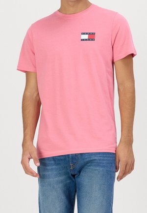 Mężczyzna w różowym T-shircie z krótkim rękawem i małym logo Tommy Jeans na klatce piersiowej oraz niebieskich dżinsach.
