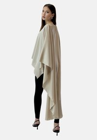 Beżowy satynowy peleryna o zwiewnym kroju, z otwartymi bokami i zamkiem z tyłu, zestawiona z czarnymi legginsami i sandałami na wysokim obcasie z paskami.