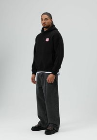 Zwarte hoodie met een roze vierkante patch met de letter "E". Gecombineerd met wijde donkergrijze jeans en zwarte schoenen met kleurrijke veters.