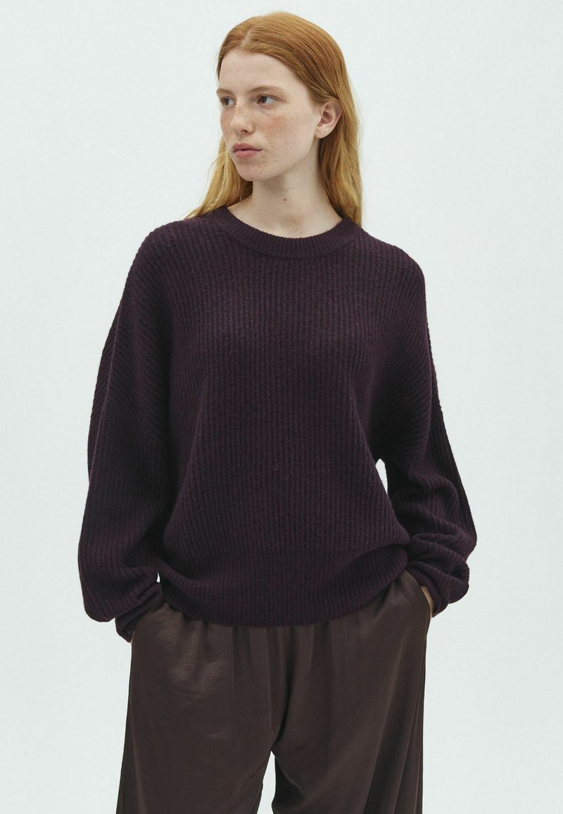 Jeune femme aux cheveux rouges portant un pull en maille violet foncé et un pantalon marron ample, debout les mains dans les poches, regardant sur le côté.