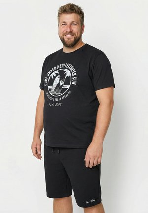 Uomo sorridente con barba che indossa una maglietta nera con grafica di tavole da surf e palma e pantaloncini neri su uno sfondo bianco semplice.