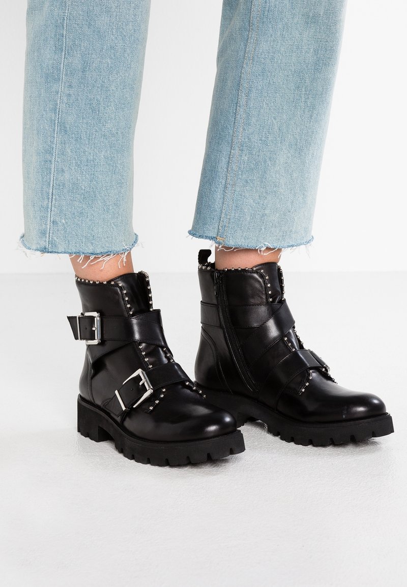Bottes en cuir noir montant à la cheville avec des clous argentés, deux sangles à boucle et des fermetures éclair latérales, portées avec un jean court bleu clair.