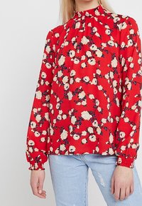 Blouse rouge à imprimé floral, col montant et manches longues avec poignets froncés. Fabriquée en tissu léger, elle présente des fleurs blanches et bleues.