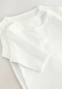 Dwa złożone białe body dla niemowląt z zapięciami na napy na ramionach i z przodu, leżące na jasnym tle.