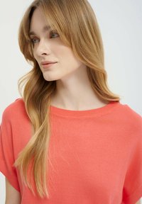 Top in corallo lavorato a maglia con scollatura ampia e maniche corte, caratterizzato da una texture morbida e una vestibilità relax, messo in risalto da capelli lisci che scivolano sulla spalla.