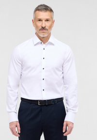 Witte langmouw shirt met een gestructureerde kraag, met donkere knopen. Draagt bij donkere broek en een zwarte geweven riem.